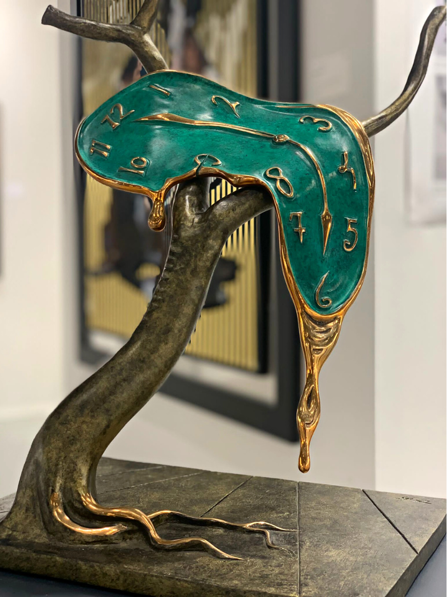 Cette image représente une sculpture de l'artiste Salvador Dalí intitulée "Profil du Temps" Réalisée en bronze à la cire perdue, elle représente une horloge fondue sur un arbres à deux branches, aux couleurs vertes et dorées avec un socle. Elle mesure 51 cm et est exposée à la Galerie Montmartre à Paris, France.
